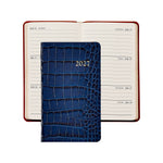 A 2027 daily planner with a dark blue crocodile-embossed cover, shown in front of an open matching planner with cream-colored pages and red-brown covers, on a white background.
