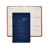 A 2027 daily planner with a dark blue crocodile-embossed cover, shown in front of an open matching planner with cream-colored pages and red-brown covers, on a white background.